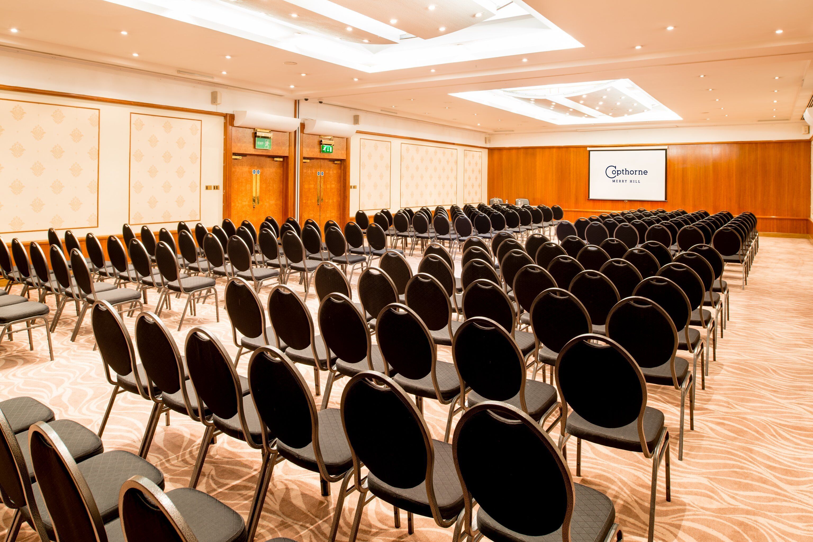 A conference room with rows of chairs, a projection screen, and a wooden wall with a patterned design.