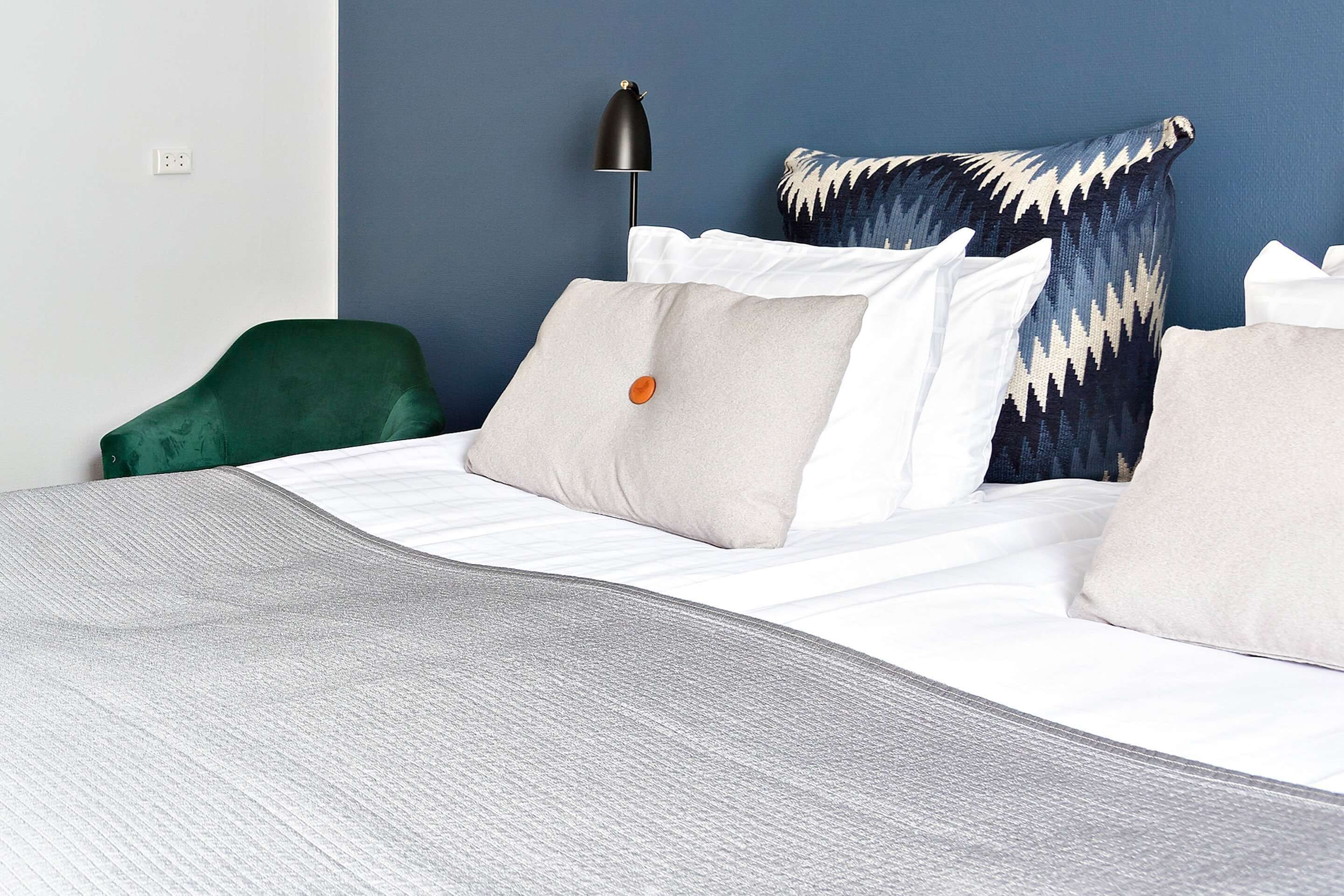 A neatly made bed with white linens and multiple pillows, a green chair, and a black floor lamp.