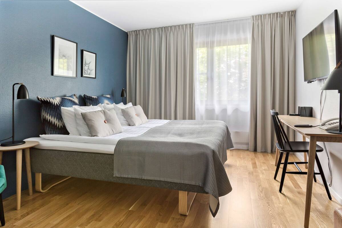 Una habitación de hotel moderna con una cama grande, un escritorio con silla, un televisor y una ventana con cortinas.
