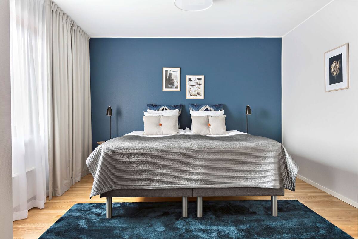 Un dormitorio con una cama, dos mesitas de noche, una pared azul y una ventana con cortinas.