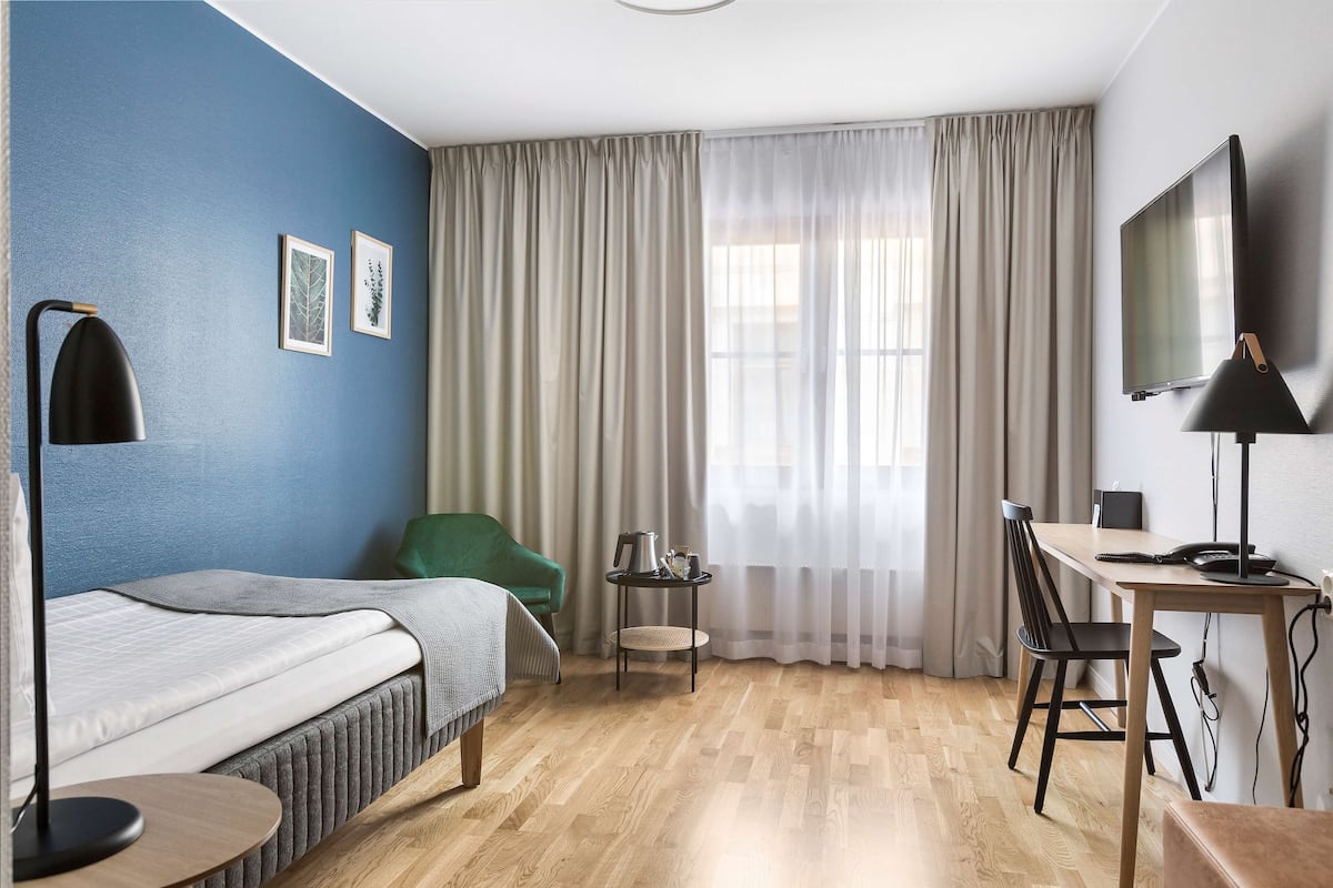 Habitación de hotel con cama, escritorio, silla, televisor y ventana con cortinas.