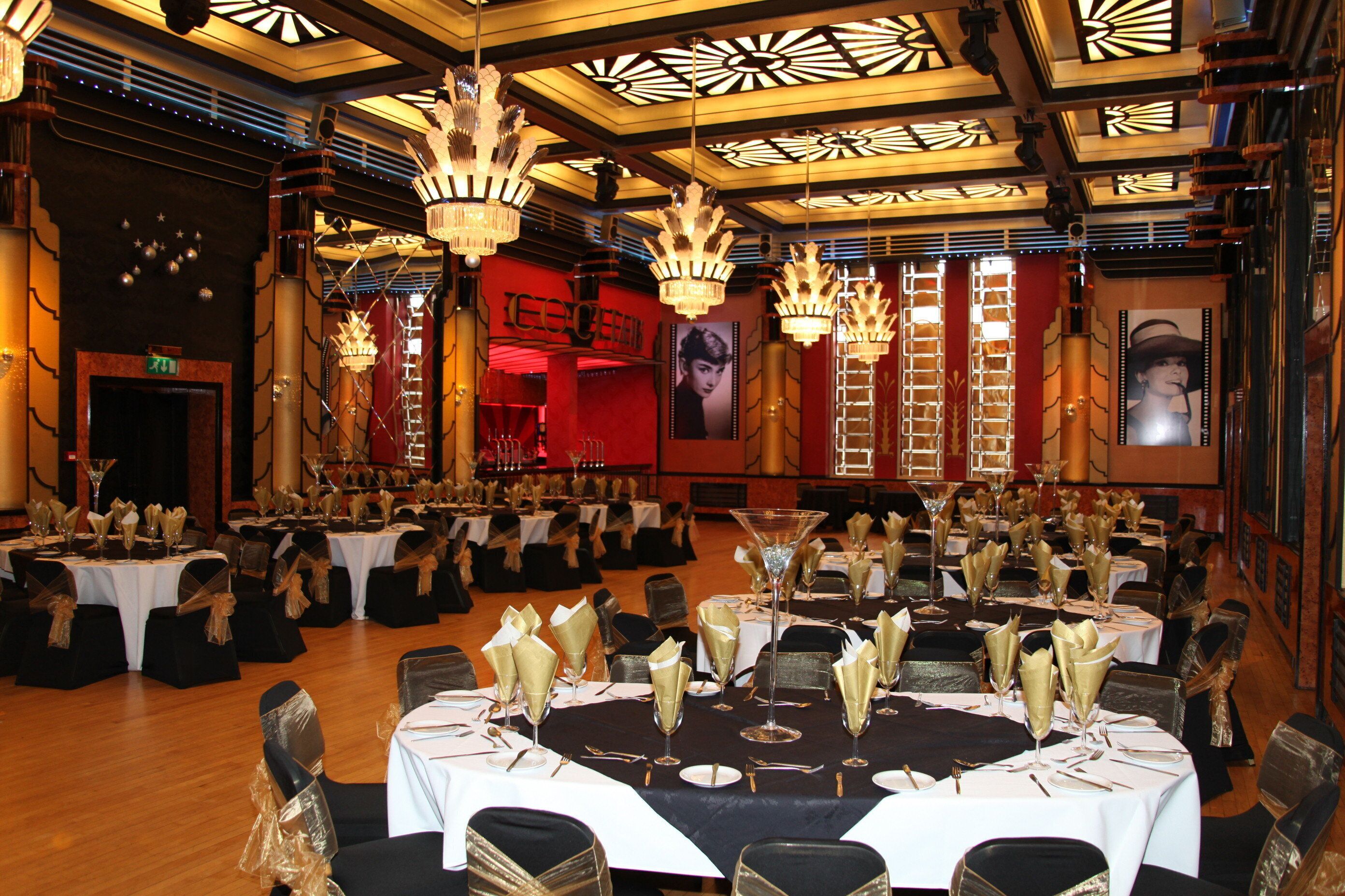 A banquet hall with round tables set for a formal event, featuring chandeliers and a red wall with a poster.