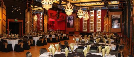 A banquet hall with round tables set for a formal event, featuring chandeliers and a red wall with a poster.