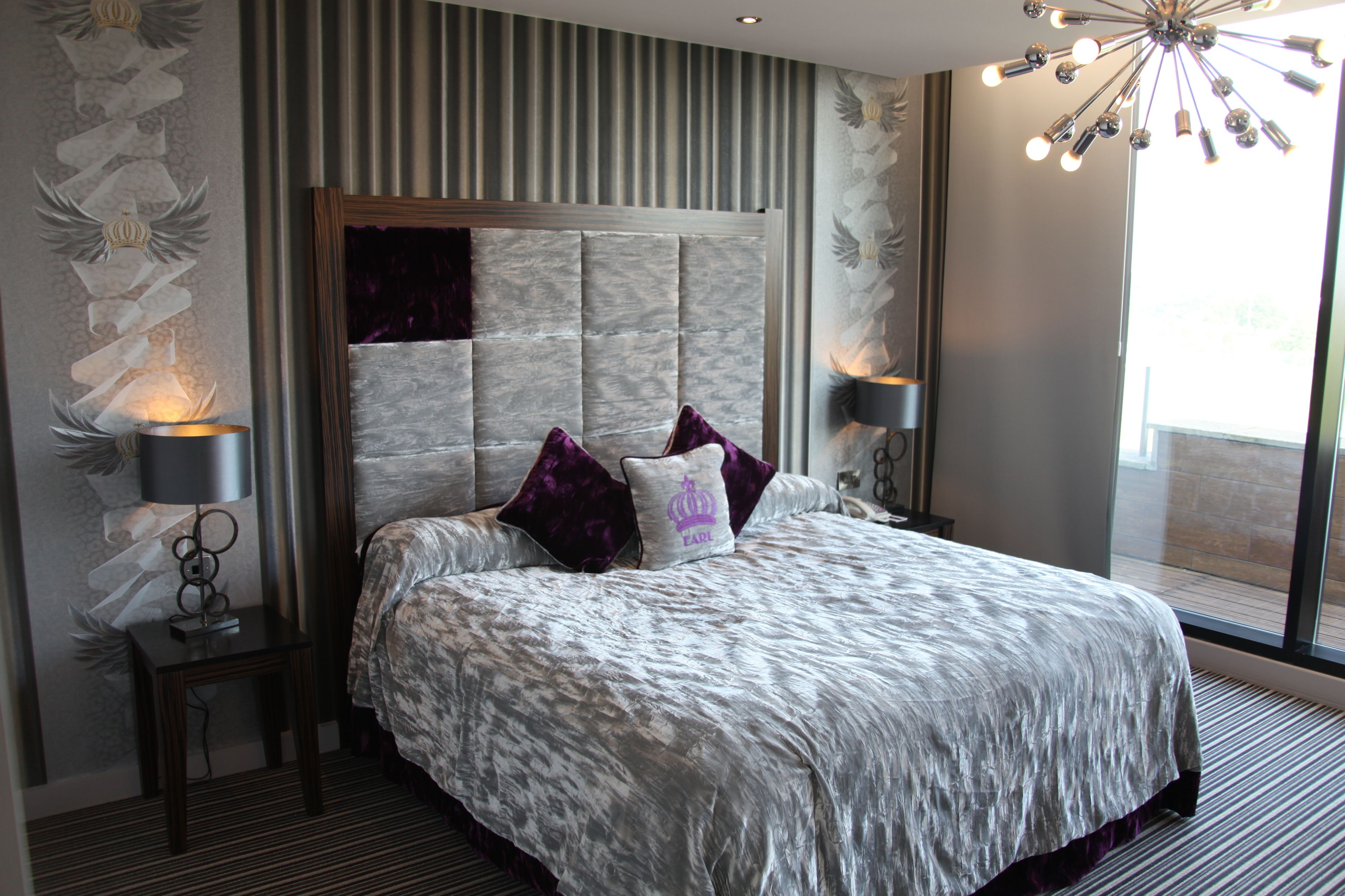 A modern hotel room with a large bed, a headboard with vertical lines, two bedside lamps, and a large window with a view.