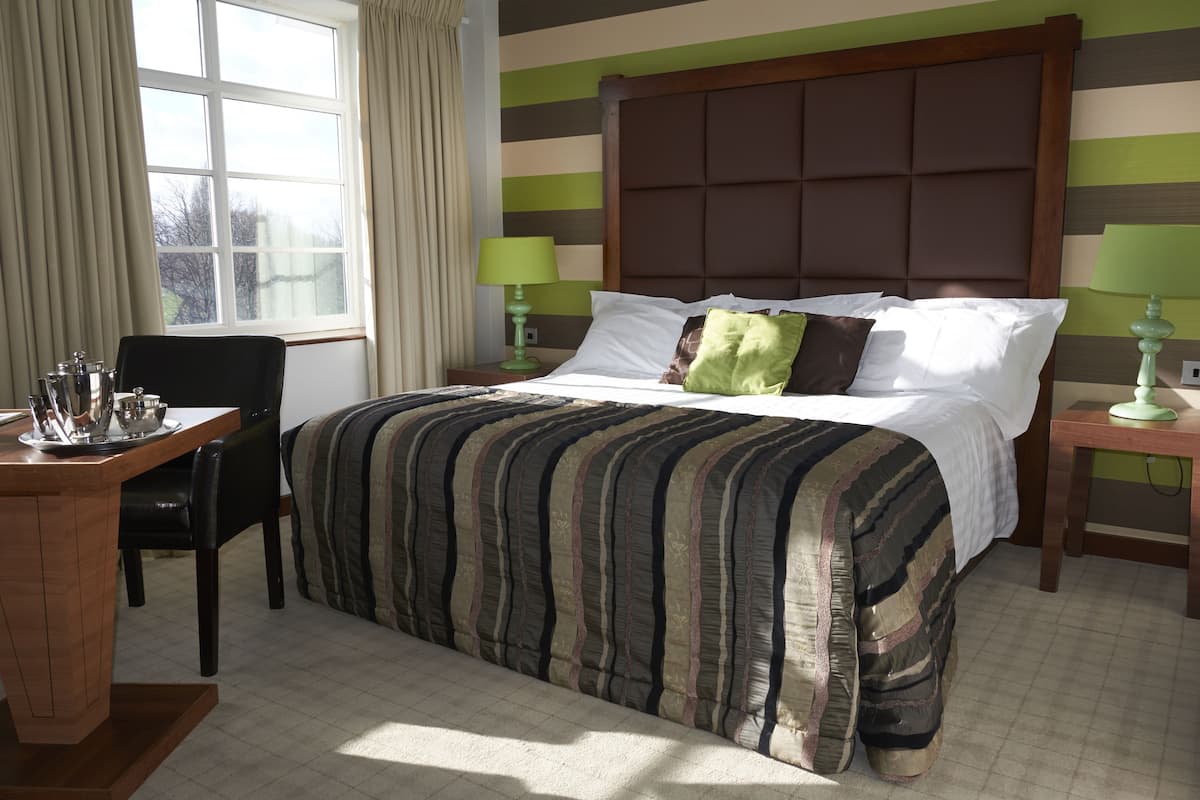 Een hotelkamer met een groot bed, gestreepte wanden, een houten hoofdbord, twee nachtkastjes met lampen, een bureau met stoel en een raam met gordijnen.