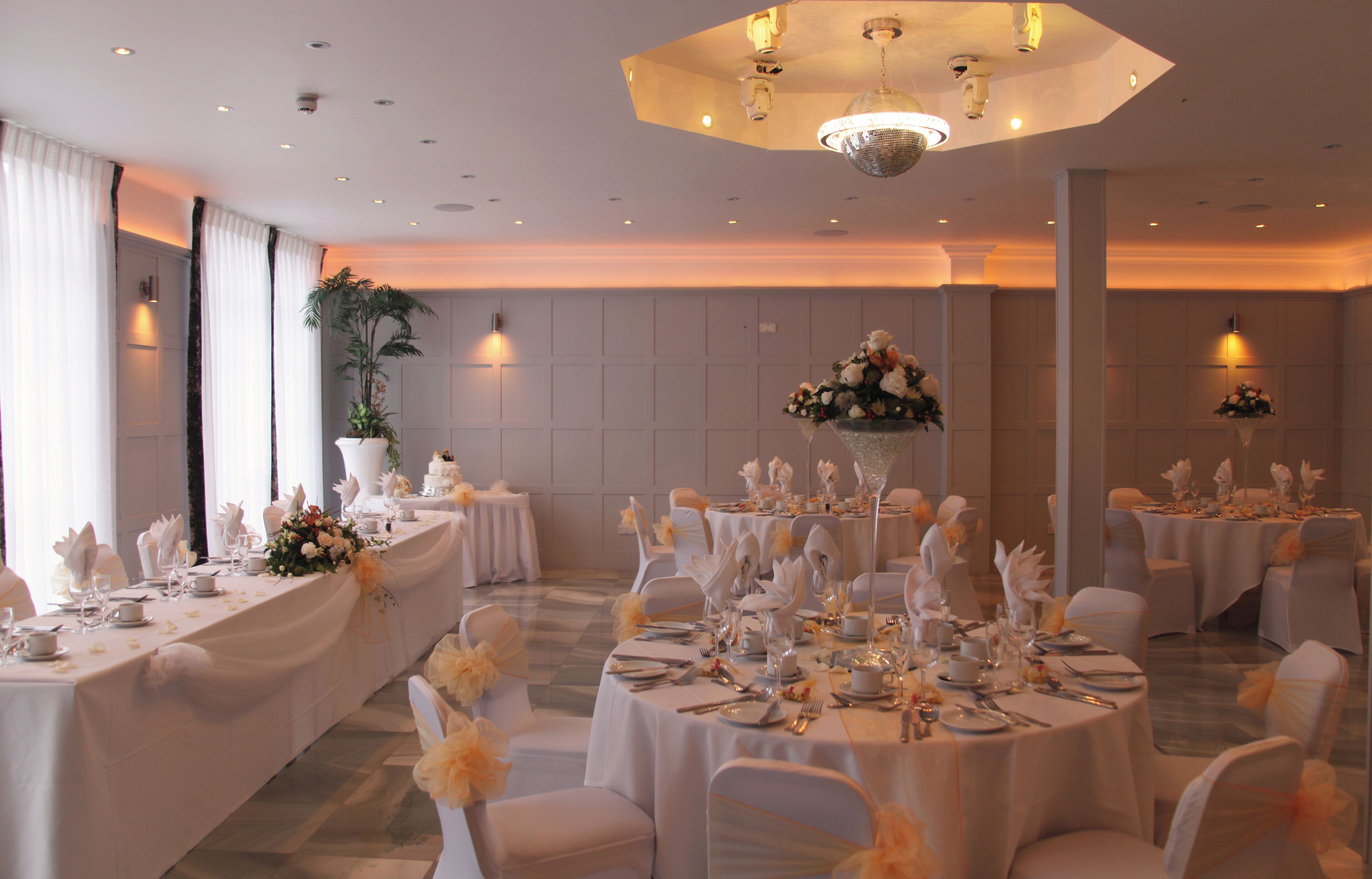 A banquet hall with round tables set for a formal event, featuring white tablecloths, floral centerpieces, and neatly arranged tableware.