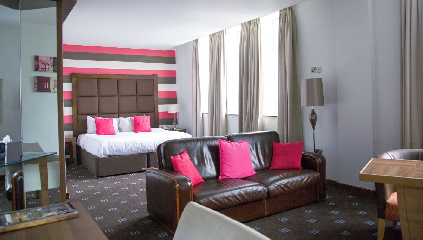 A hotel room with a bed, a leather sofa, a glass table, and a lamp.