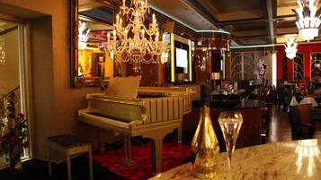 A grand piano, a chandelier, and a bottle of champagne on a marble table in a luxurious room with a red carpet.