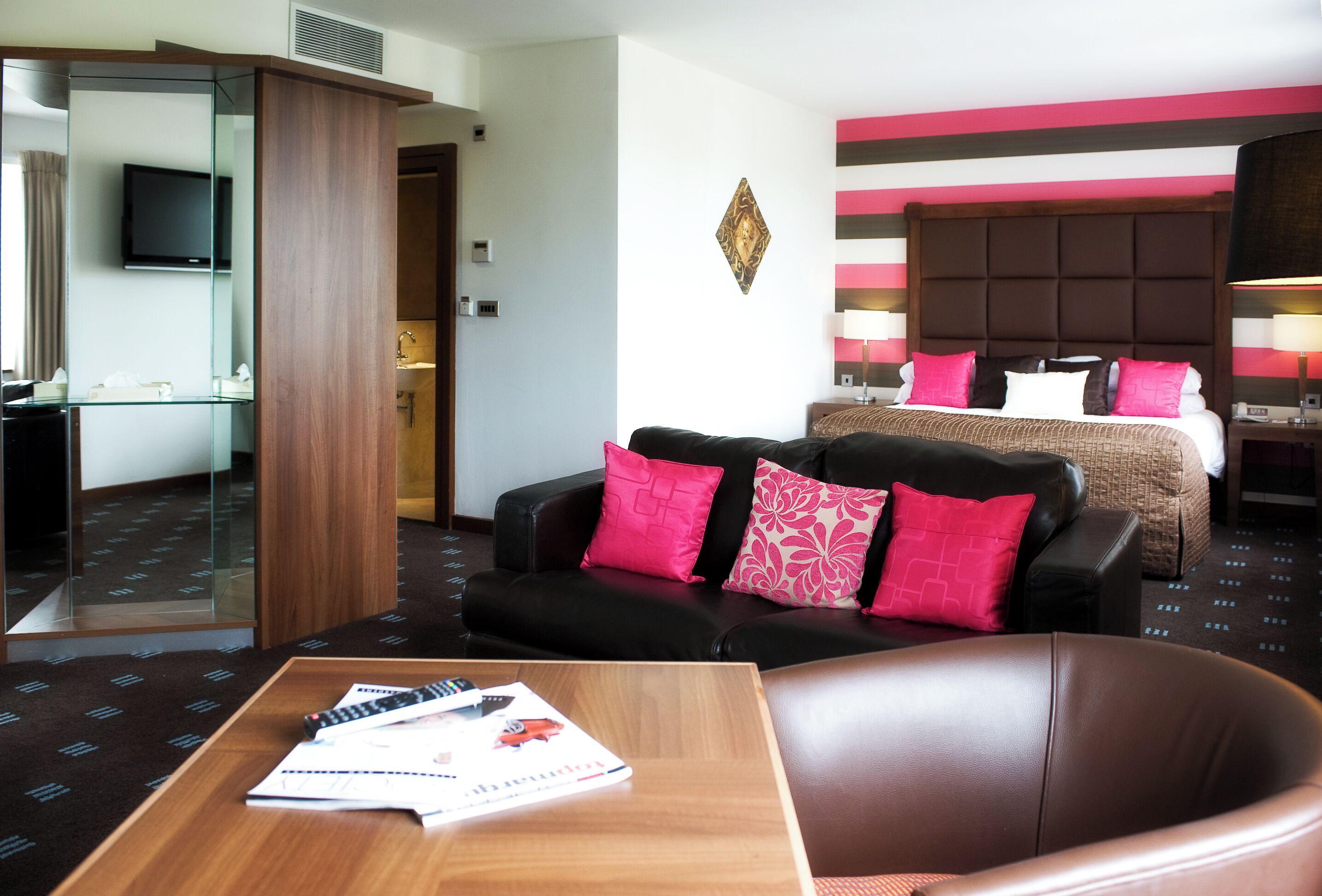 Une chambre d’hôtel avec un lit, un canapé en cuir noir, une table basse en bois sur laquelle se trouvent des papiers, une télévision et une salle de bain visible à travers une paroi en verre.