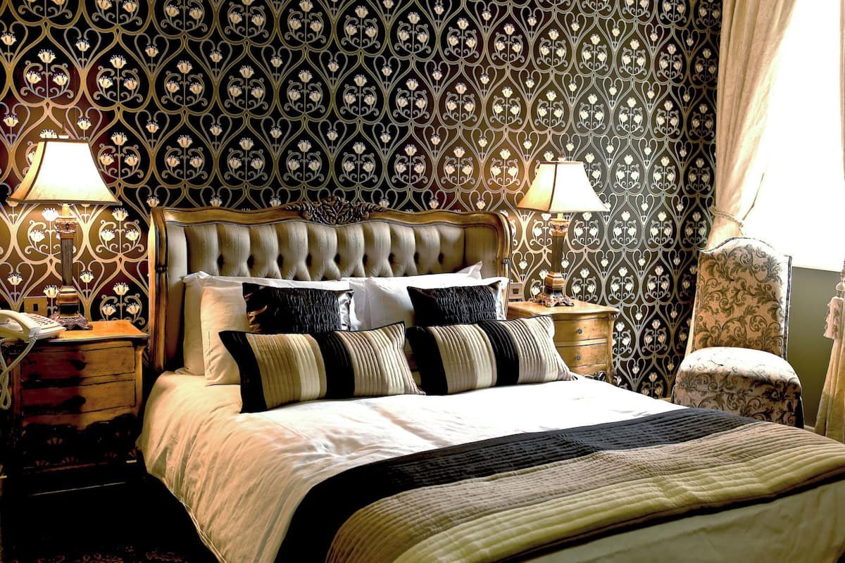 Een slaapkamer met een gebutste hoofdbord, een bed met gestreept beddengoed, twee bedlampen, een houten nachtkastje en behang met een patroon.