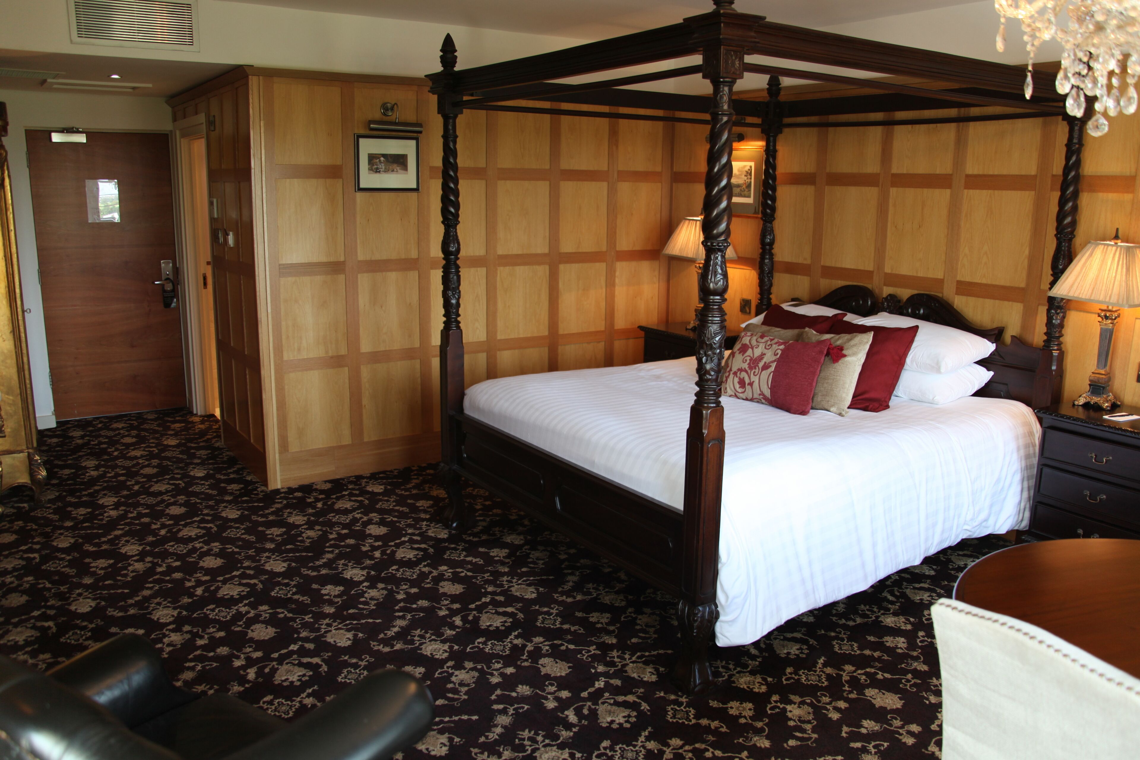 A four-poster bed with a wooden frame and white bedding, a wooden wardrobe, a desk with a lamp, and a patterned carpet.