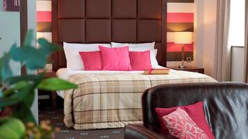 A hotel room with a large bed, a headboard, a bedside table with a lamp, a black leather chair, and a potted plant.