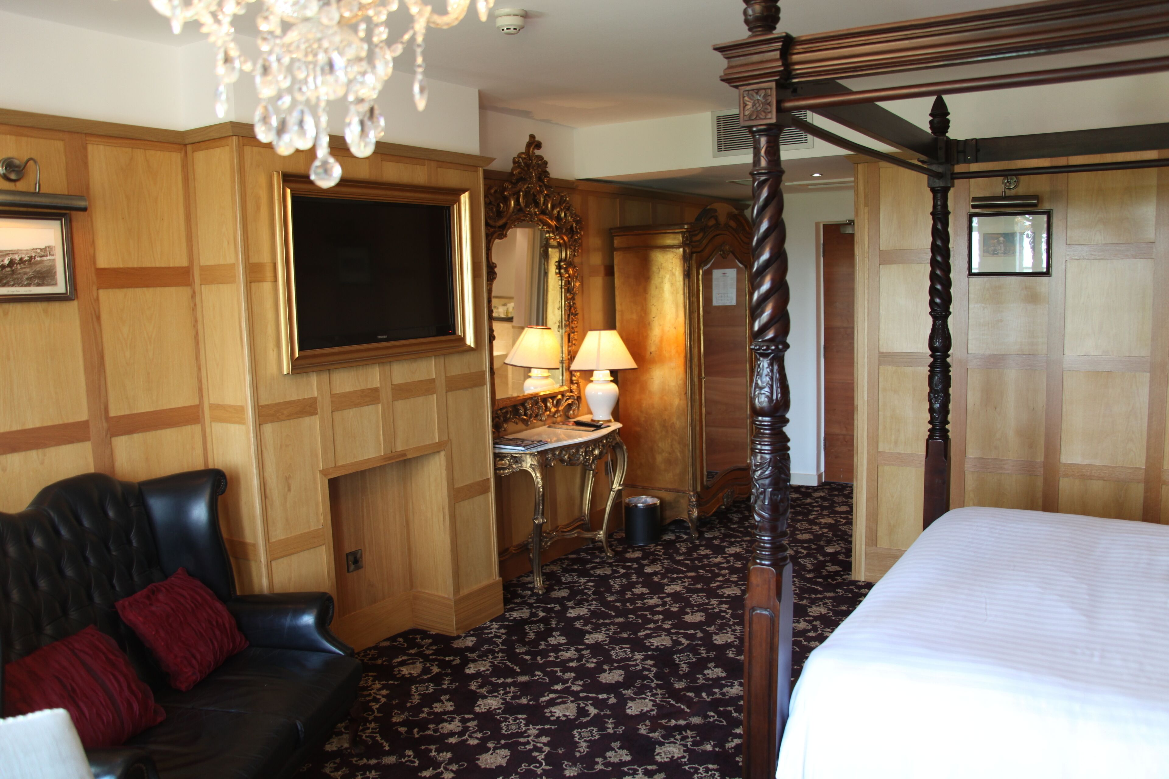 Habitación de hotel con cama con dosel, sofá de cuero, candelabro, televisor y pared de paneles de madera.