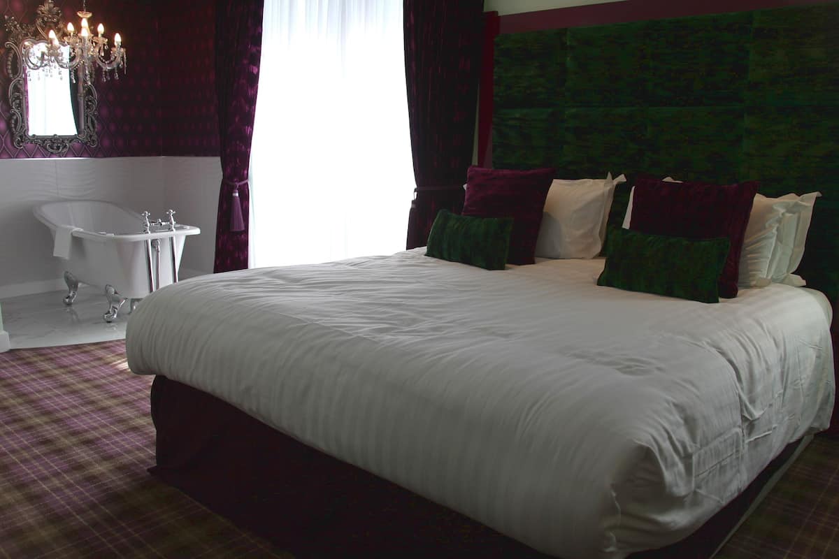 Een groot bed met witte lakens, een groen hoofdeinde en meerdere kussens. Er is een wit bad en een spiegel erboven. De kamer heeft een geruite vloer en een kroonluchter.
