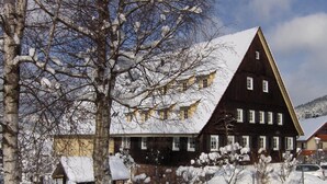 A large, dark wooden building with multiple windows and a sloped roof, surrounded by snow-covered trees and a fence. - Gutshof Hotel Waldknechtshof (Baiersbronn)