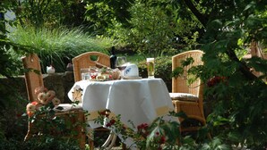 An outdoor dining area with wicker chairs and a table set for two, surrounded by lush greenery.