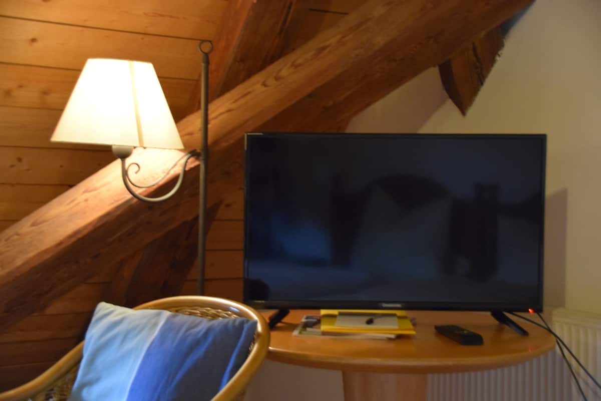 A room with a television on a wooden table, a lamp on the wall, and a chair with a cushion.