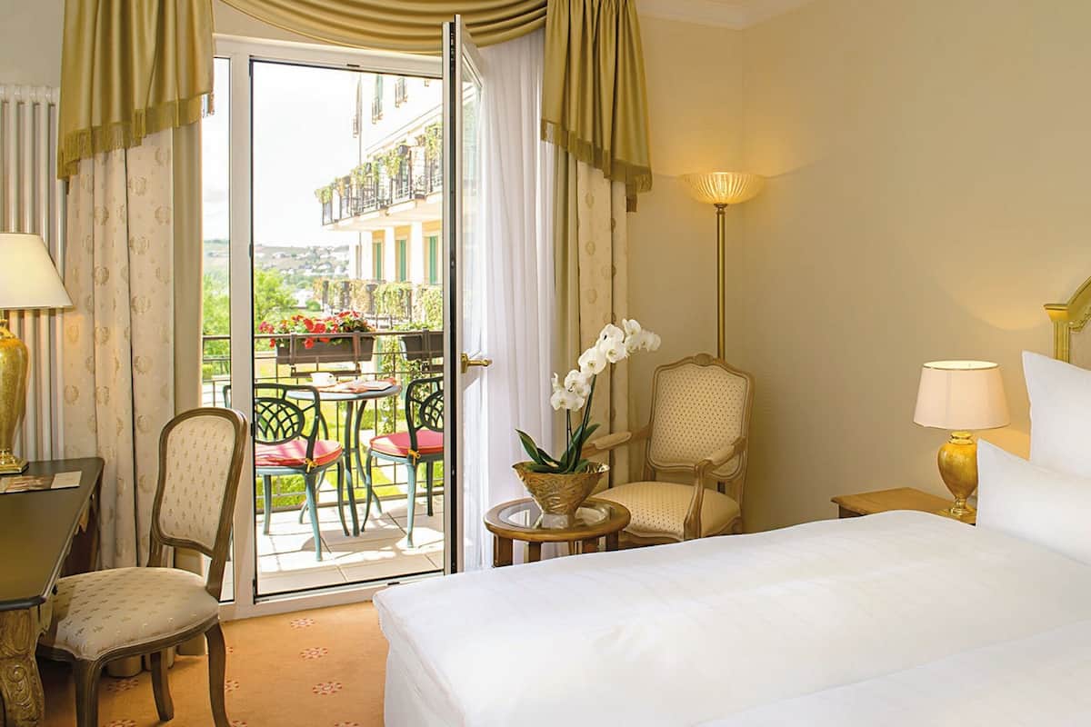 Une chambre d’hôtel avec un lit, une chaise, une petite table et une lampe. La fenêtre donne sur un balcon aménagé avec des meubles d’extérieur et des plantes.