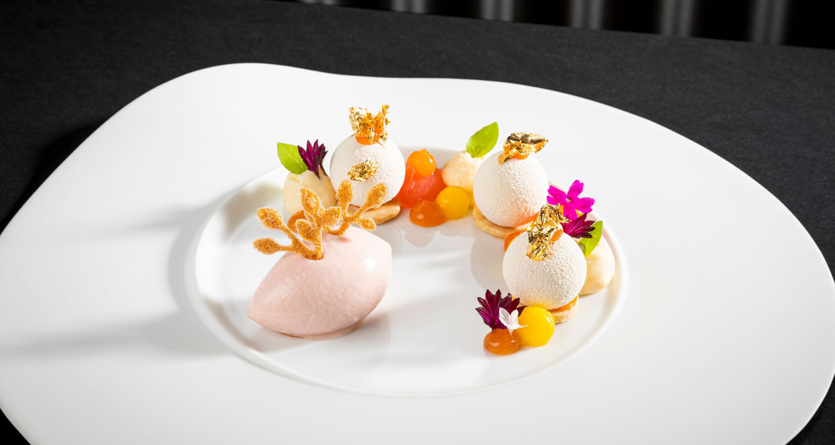 Un plato blanco con tres postres esféricos, cada uno coronado con hojas de oro y rodeado de adornos coloridos.