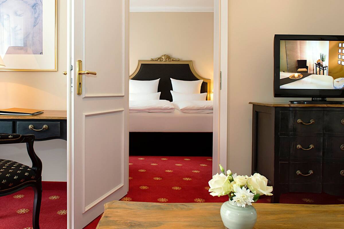 Une chambre d’hôtel avec un lit, un bureau avec une lampe, une chaise, une télévision et un vase avec des fleurs.