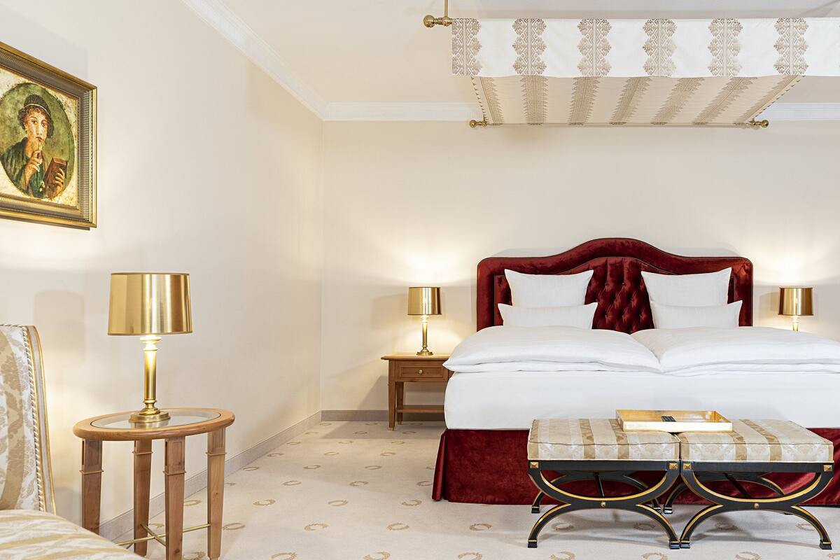Une chambre d’hôtel avec un grand lit, deux lampes de chevet, une petite table ronde, un banc et une œuvre d’art encadrée au mur.