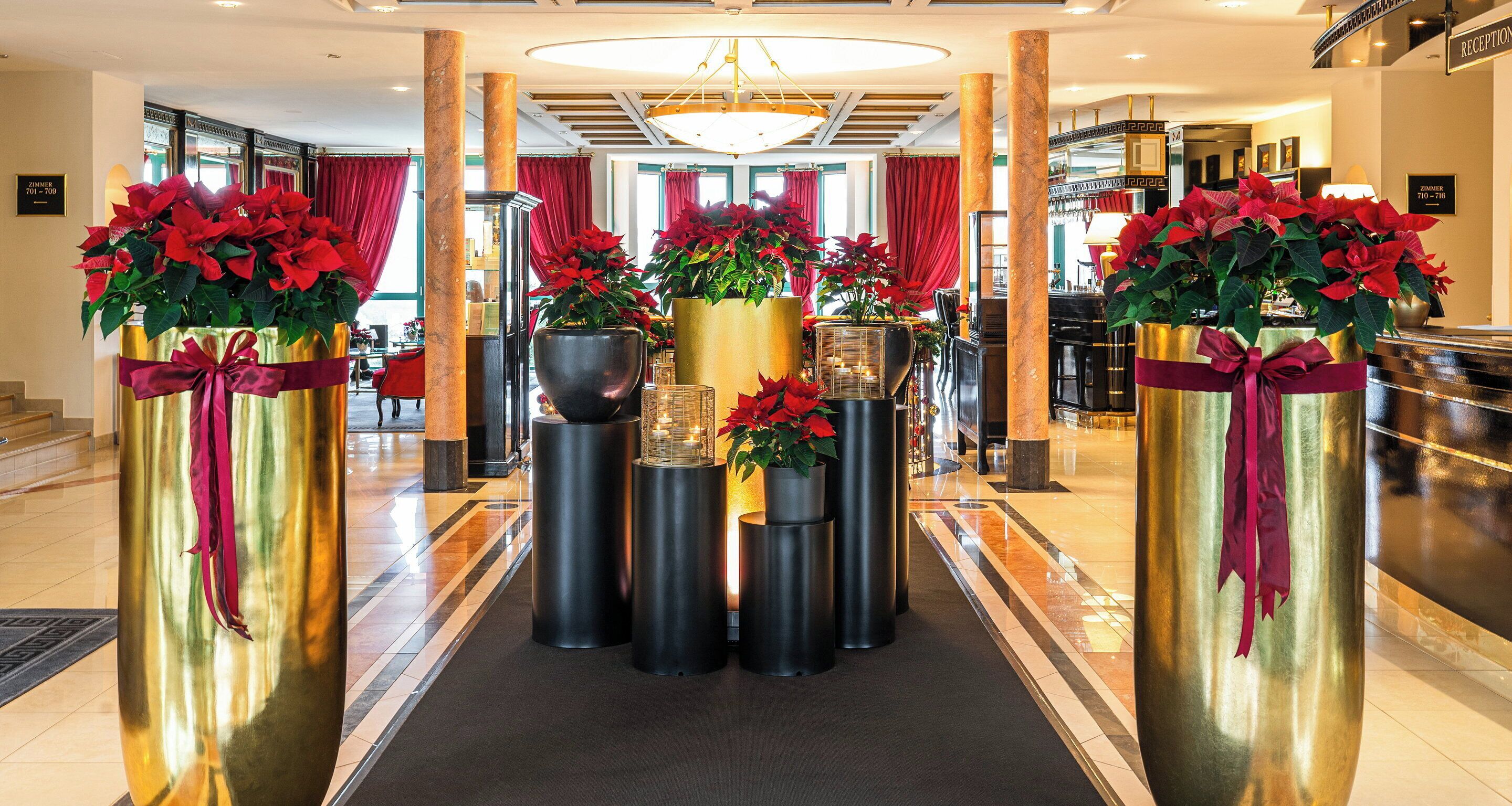 A hotel lobby with a central reception desk, large decorative vases with red flowers, and a chandelier.