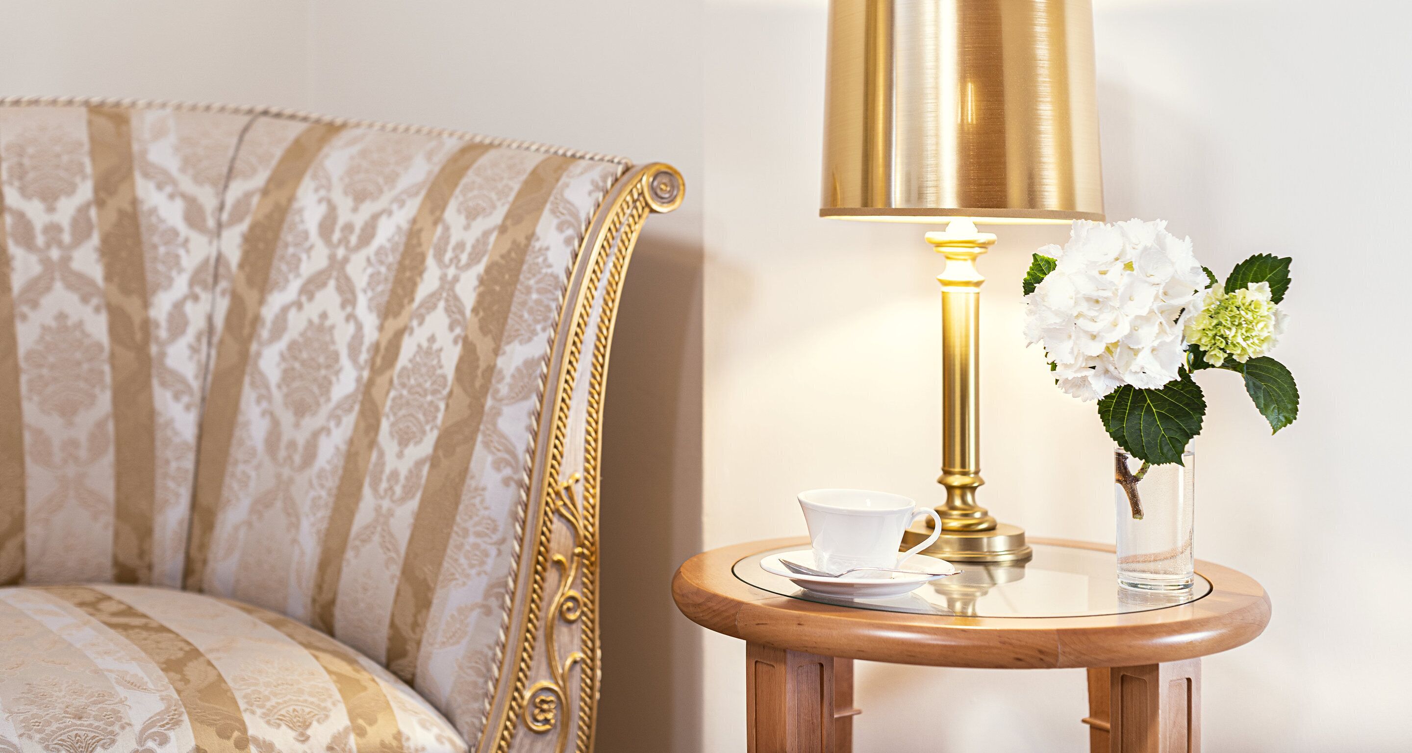 Ett rum med en mönstrad soffa, ett litet träbord med en vit kopp och ett tillhörande fat, en guldfärgad lampa och en vas med vita blommor.