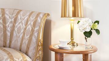 Ett rum med en mönstrad soffa, ett litet träbord med en vit kopp och ett tillhörande fat, en guldfärgad lampa och en vas med vita blommor.