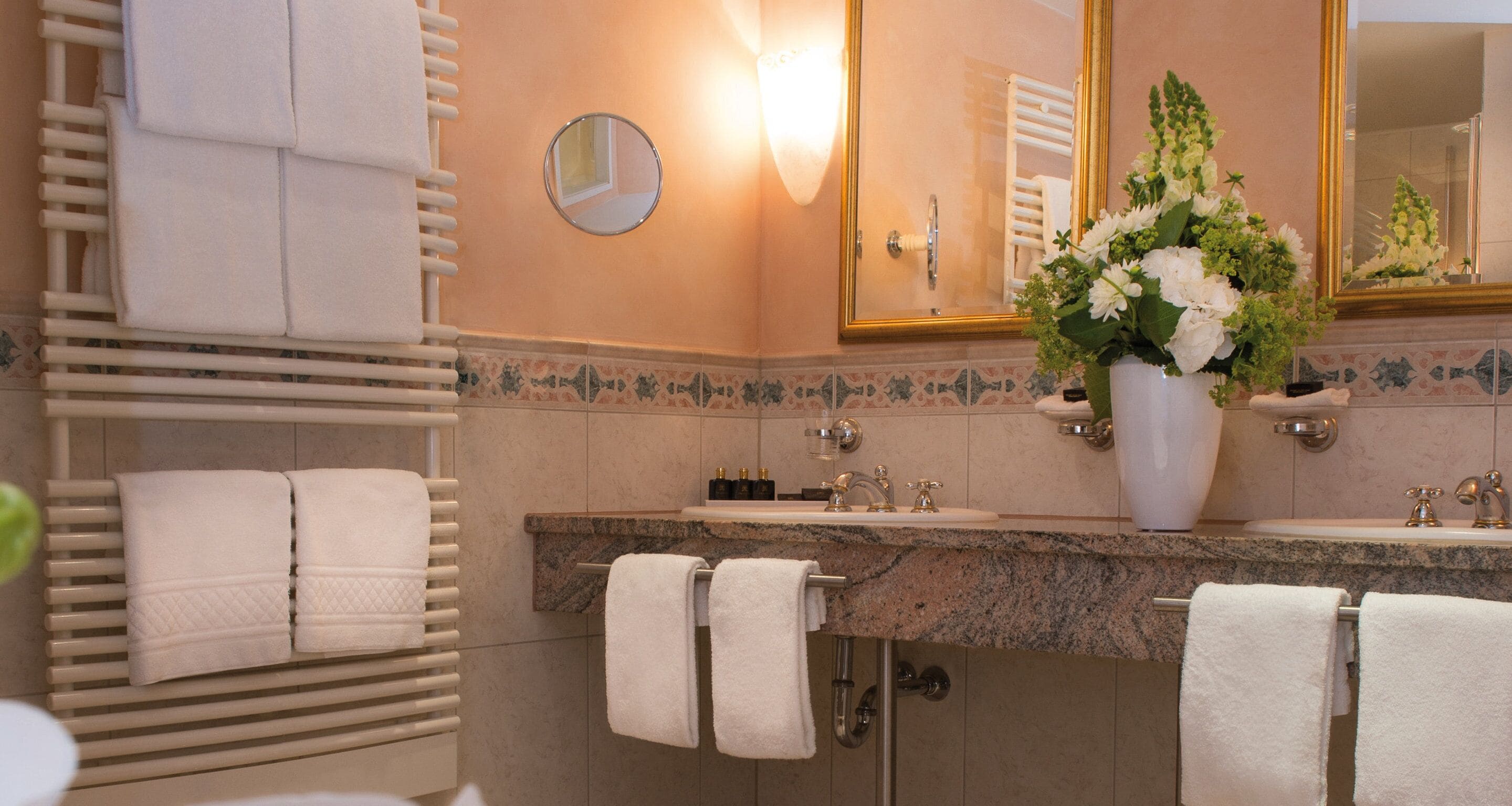 Un baño con encimera de granito, toallas blancas, un espejo redondo y un jarrón con flores.