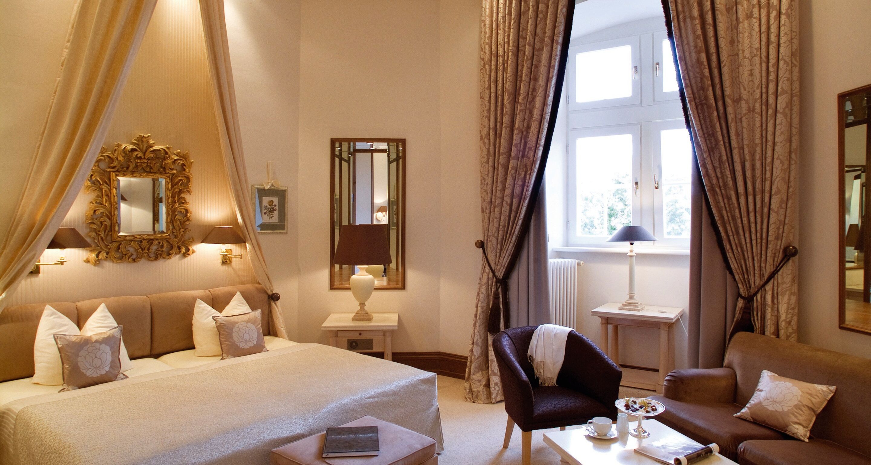 A hotel room with a large bed, a chair, a table with a cup and saucer, and a window with curtains.