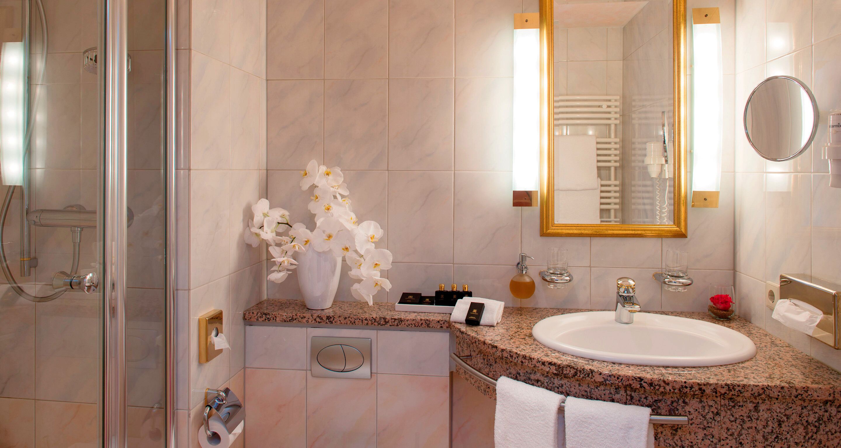 A bathroom with a glass shower enclosure, a granite countertop, a white sink, a mirror, and a vase with white flowers.
