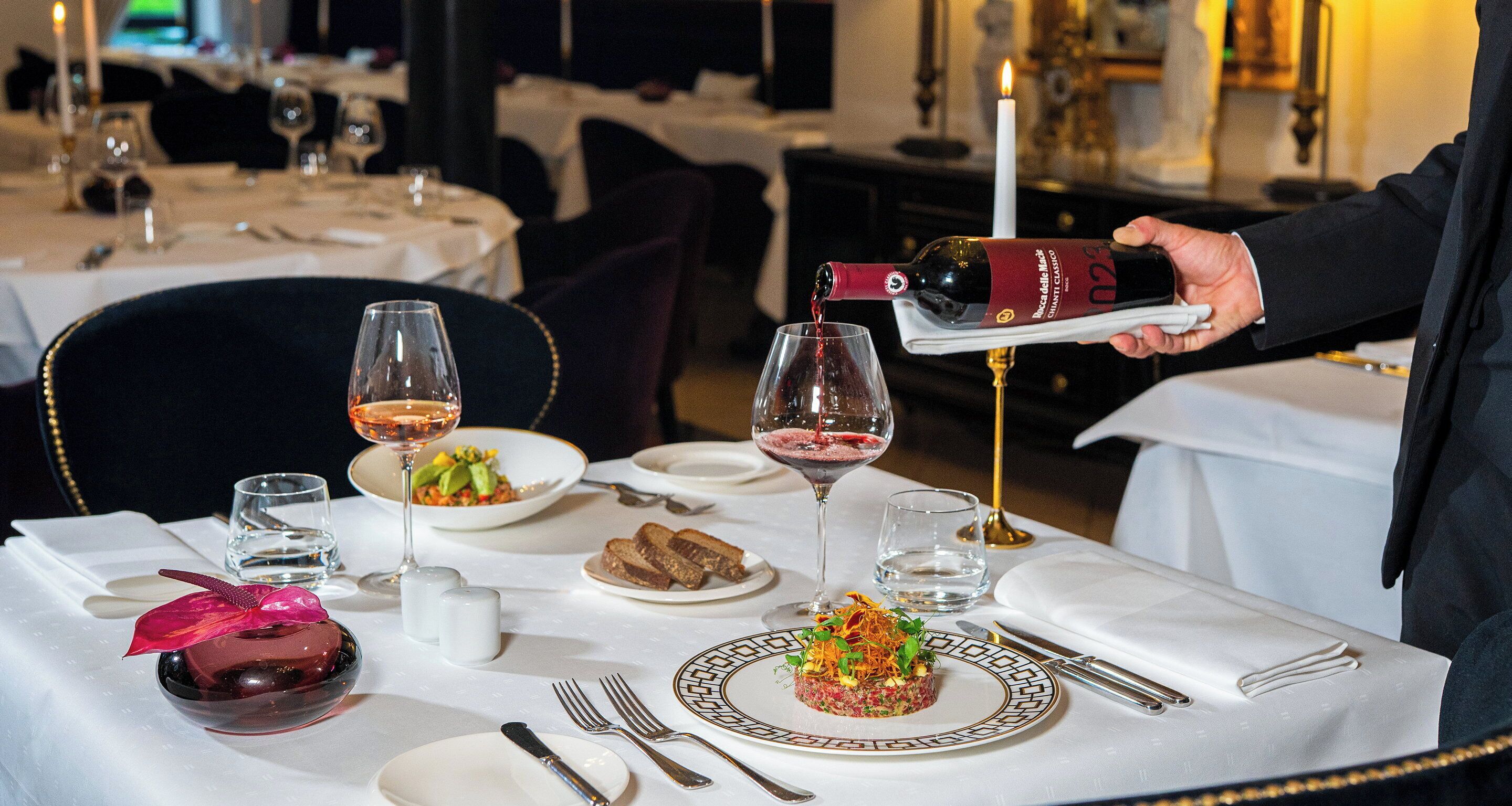 Una mesa de comedor dispuesta con platos, vasos, cubiertos y una botella vertiéndose en un vaso.