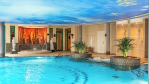 Un área de piscina interior lujosa, con un jacuzzi central, rodeado de macetas y estatuas con mosaicos.