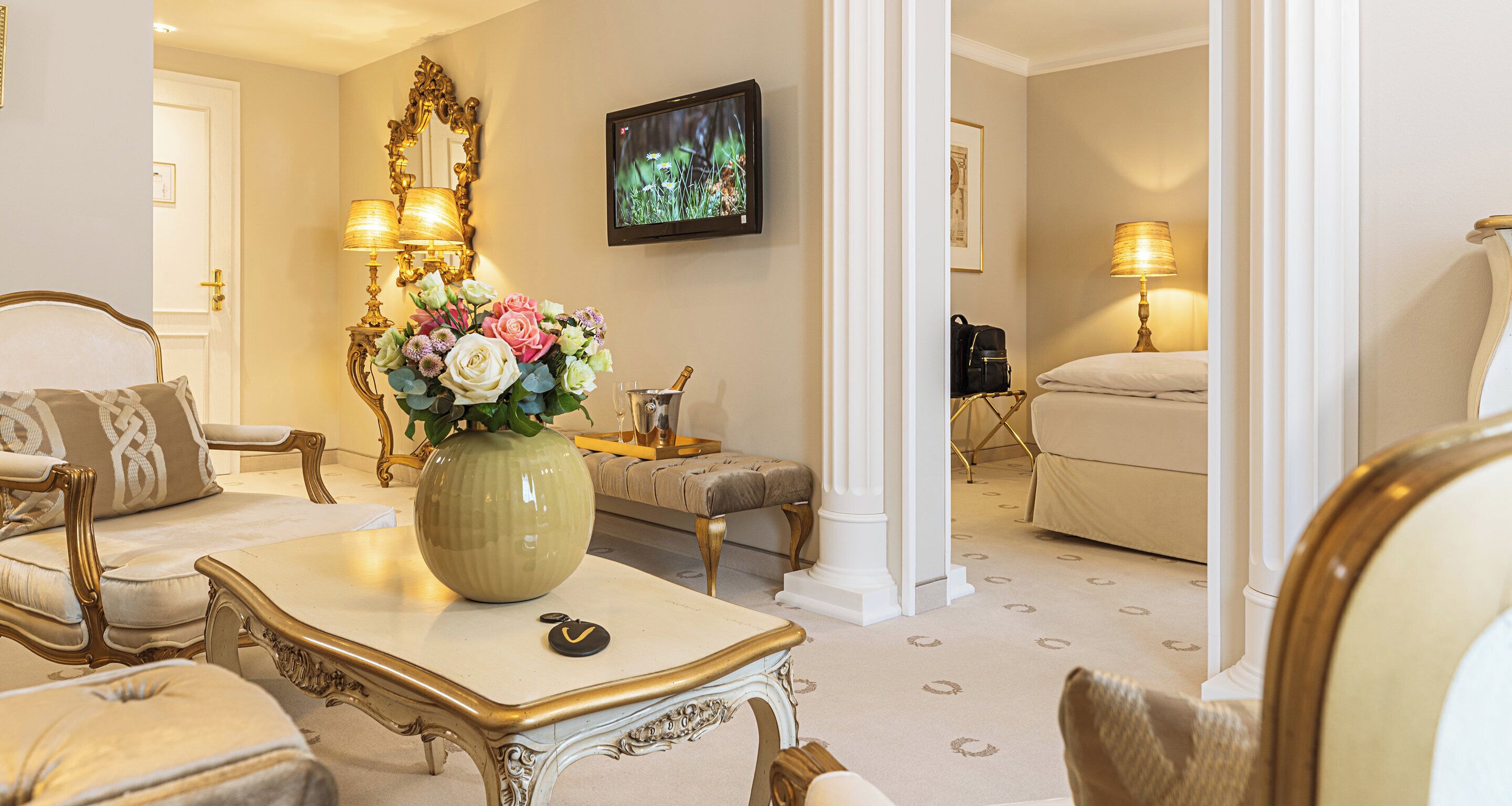 Een stijlvol ingerichte woonkamer met een grote bloemenvaas, elegant meubilair en een aan de muur bevestigde televisie.