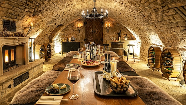 Un comedor acogedor con paredes de piedra, una larga mesa de madera dispuesta para una comida, velas y botellas de vino.