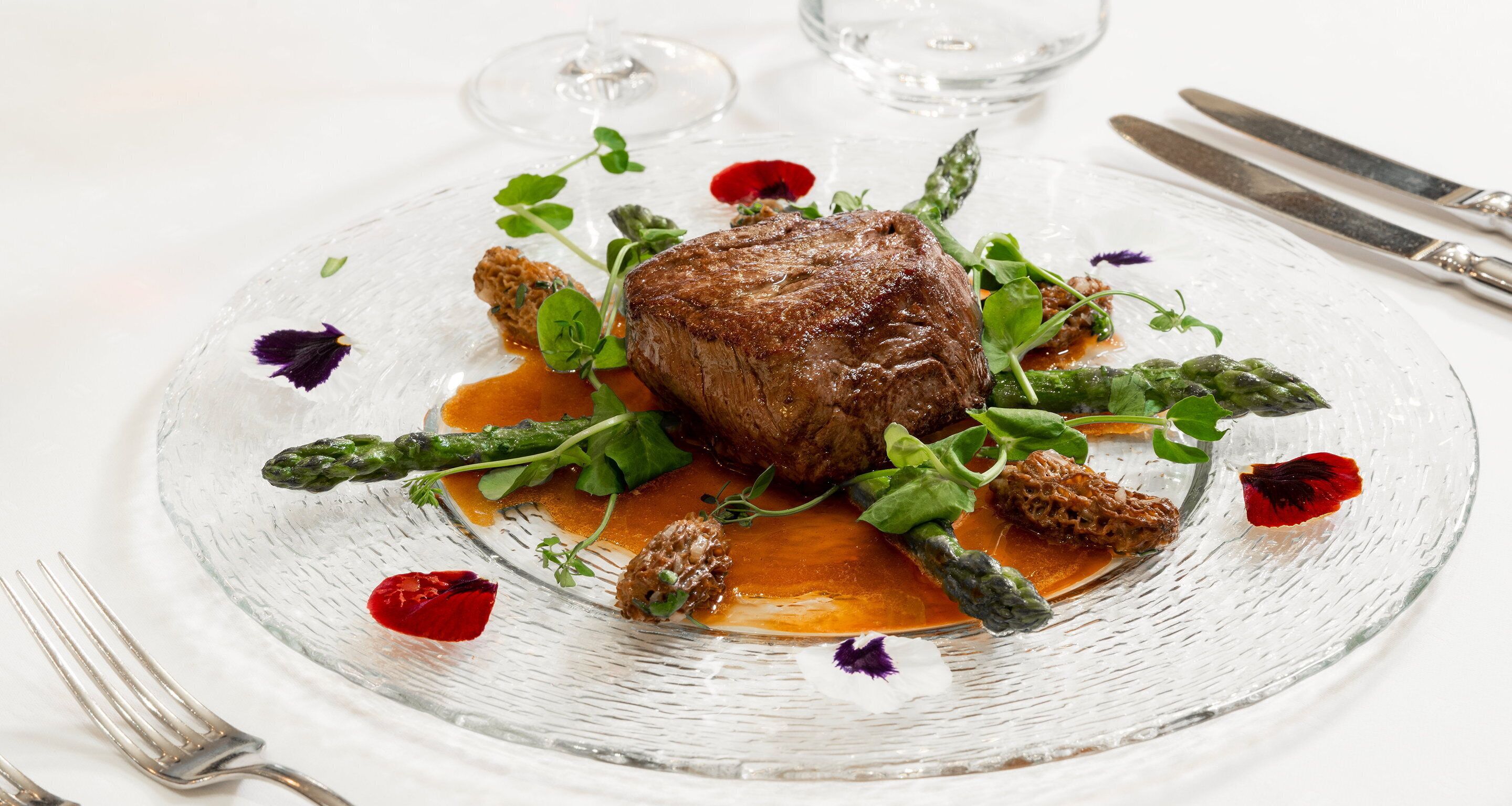 Un piatto di cibo con bistecca, asparagi e verdure, accompagnato da un bicchiere d'acqua e posate su un tavolo bianco.