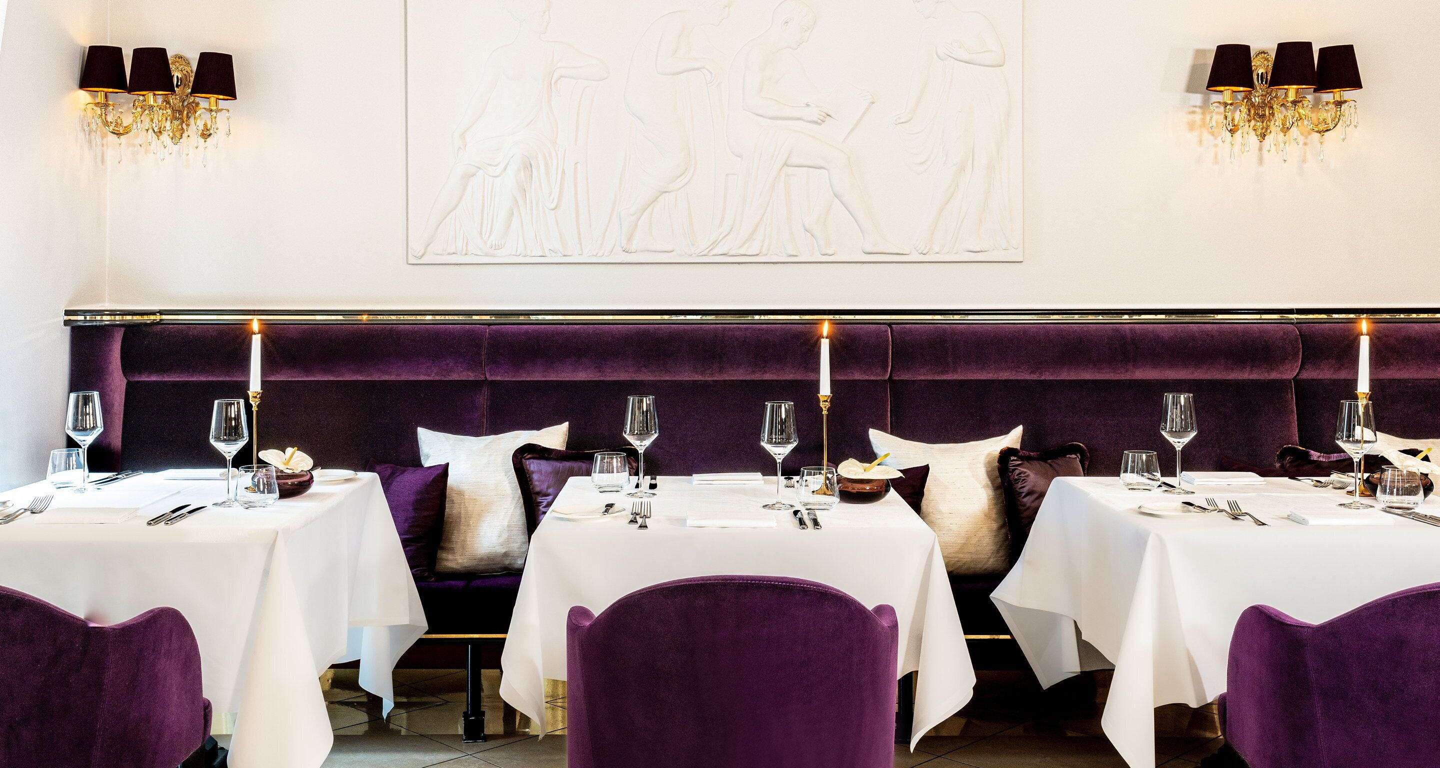 A dining area with tables set for four, each with white tablecloths, purple chairs, and candles.