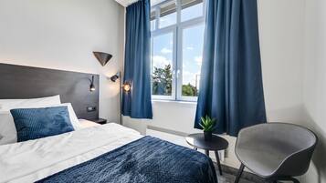 A modern hotel room with a bed, a chair, a small table, and a window with curtains.