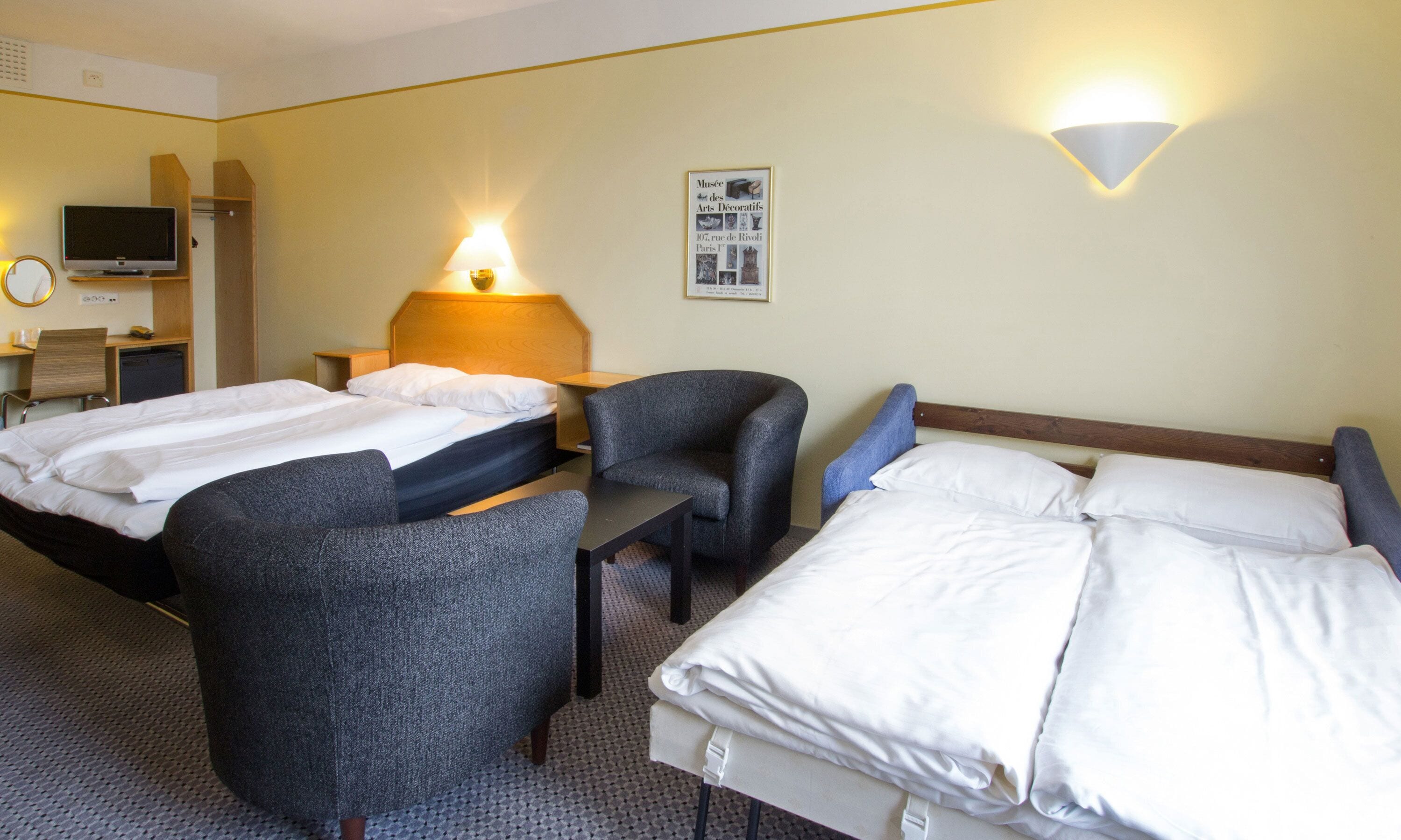 Une chambre d’hôtel avec deux lits, un bureau, une chaise, une télévision et une lampe fixée au mur.