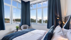 Ein Hotelzimmer mit großem Fenster, einem Bett mit weißblauer Bettwäsche, einem kleinen runden Tisch mit Pflanze und einem Sessel.