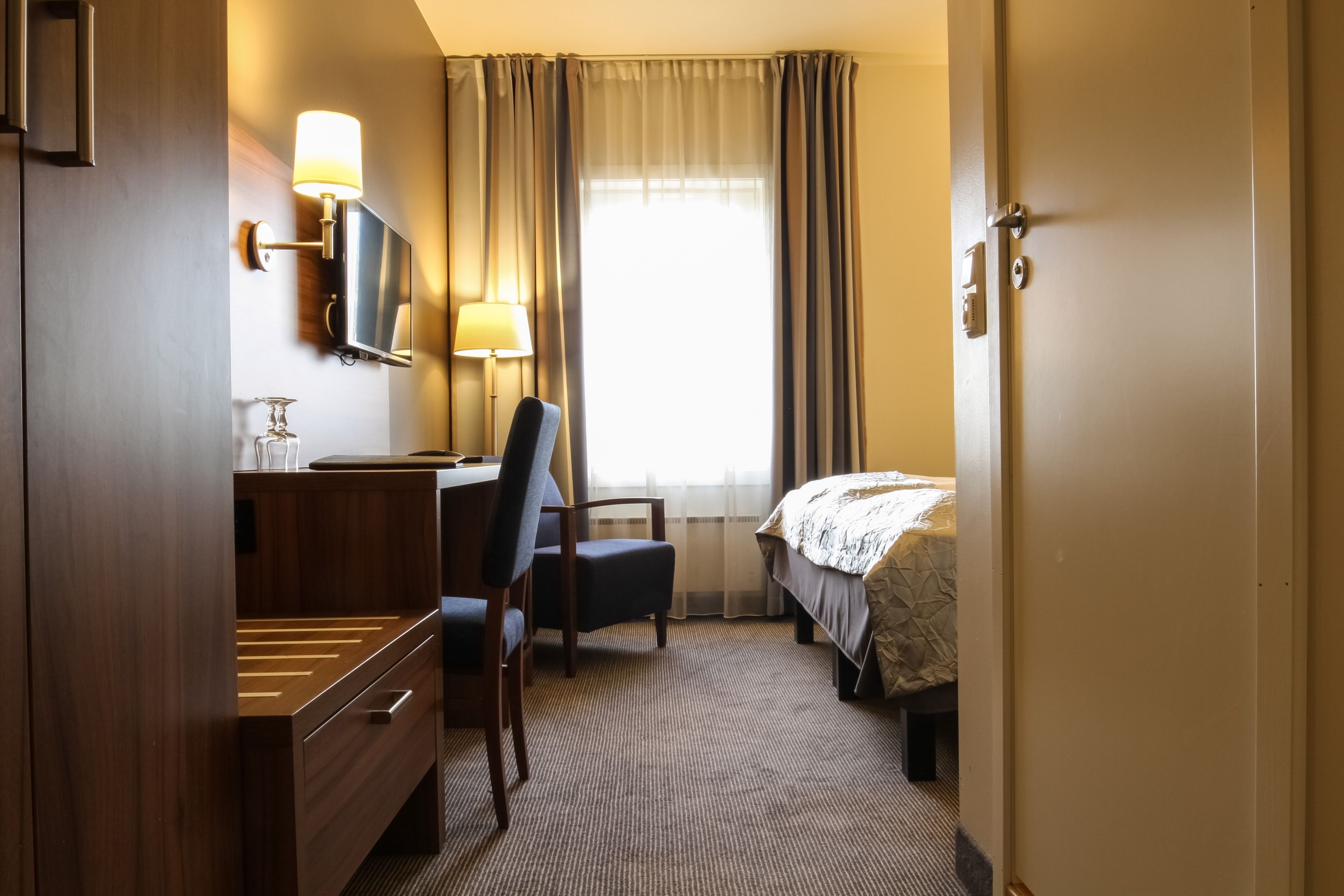A hotel room with a bed, a desk with a chair, a TV, and a lamp.