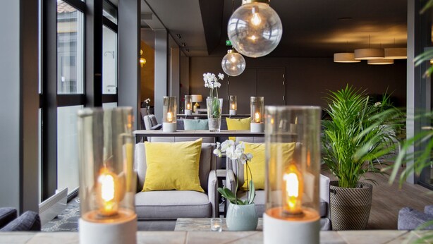 Ein modernes Interieur mit einer Sitzecke, bestehend aus einem Sofa mit gelben Kissen, einem Tisch mit Glaslaternen und einer Topfpflanze.