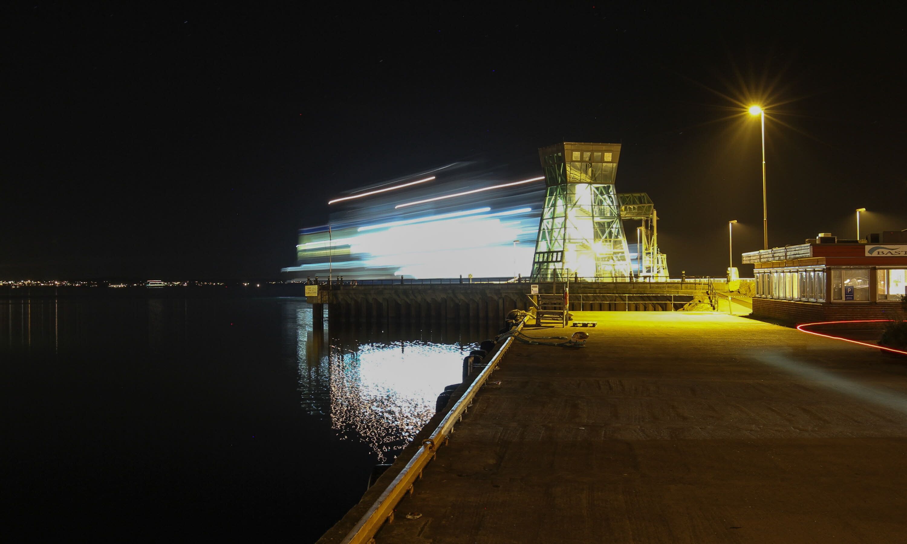 Un muelle frente al agua con una gran estructura iluminada y un edificio con la leyenda 'BAS'.