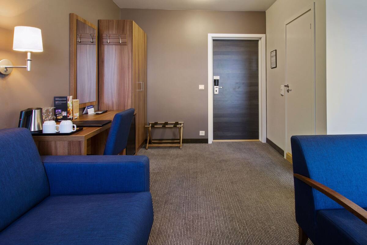 A hotel room with a desk, two blue armchairs, a small bench, a coffee maker, and a lamp.