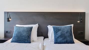 Cama doble con sábanas blancas y dos almohadas azules, cabecera oscura y dos lámparas de lectura fijadas a la pared.