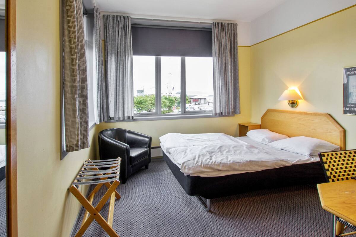 A hotel room with a bed, a chair, a small table, and a window with curtains.