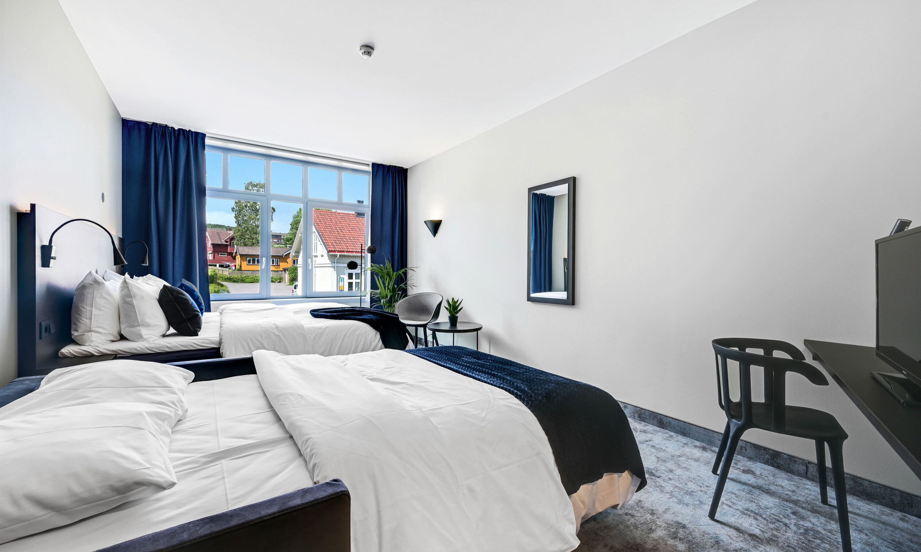 A hotel room with two beds, a desk, a chair, a small table with a plant, and a window with curtains.