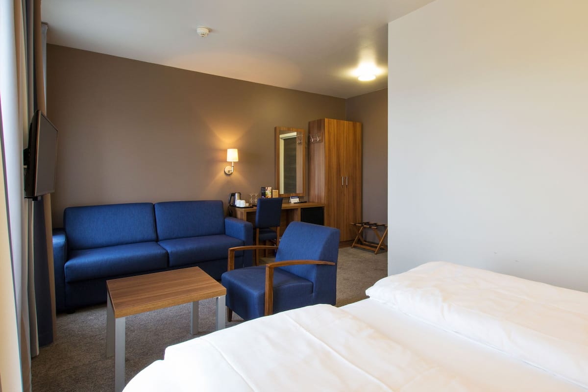 A hotel room with a blue sofa, a small wooden table, a desk with a chair, a wardrobe, and a bed.