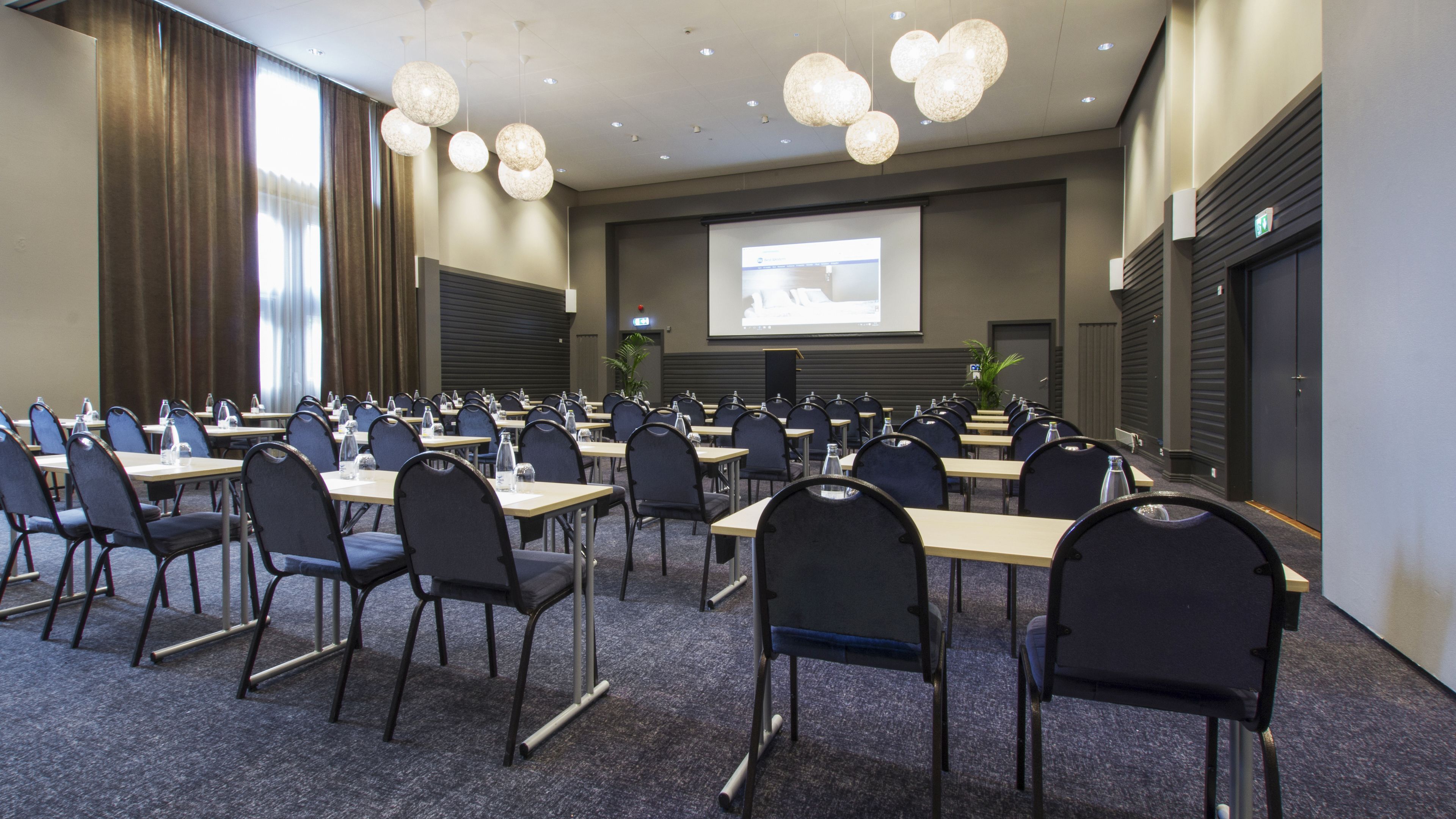 A conference room with rows of tables and chairs, a projector screen, and hanging spherical light fixtures.