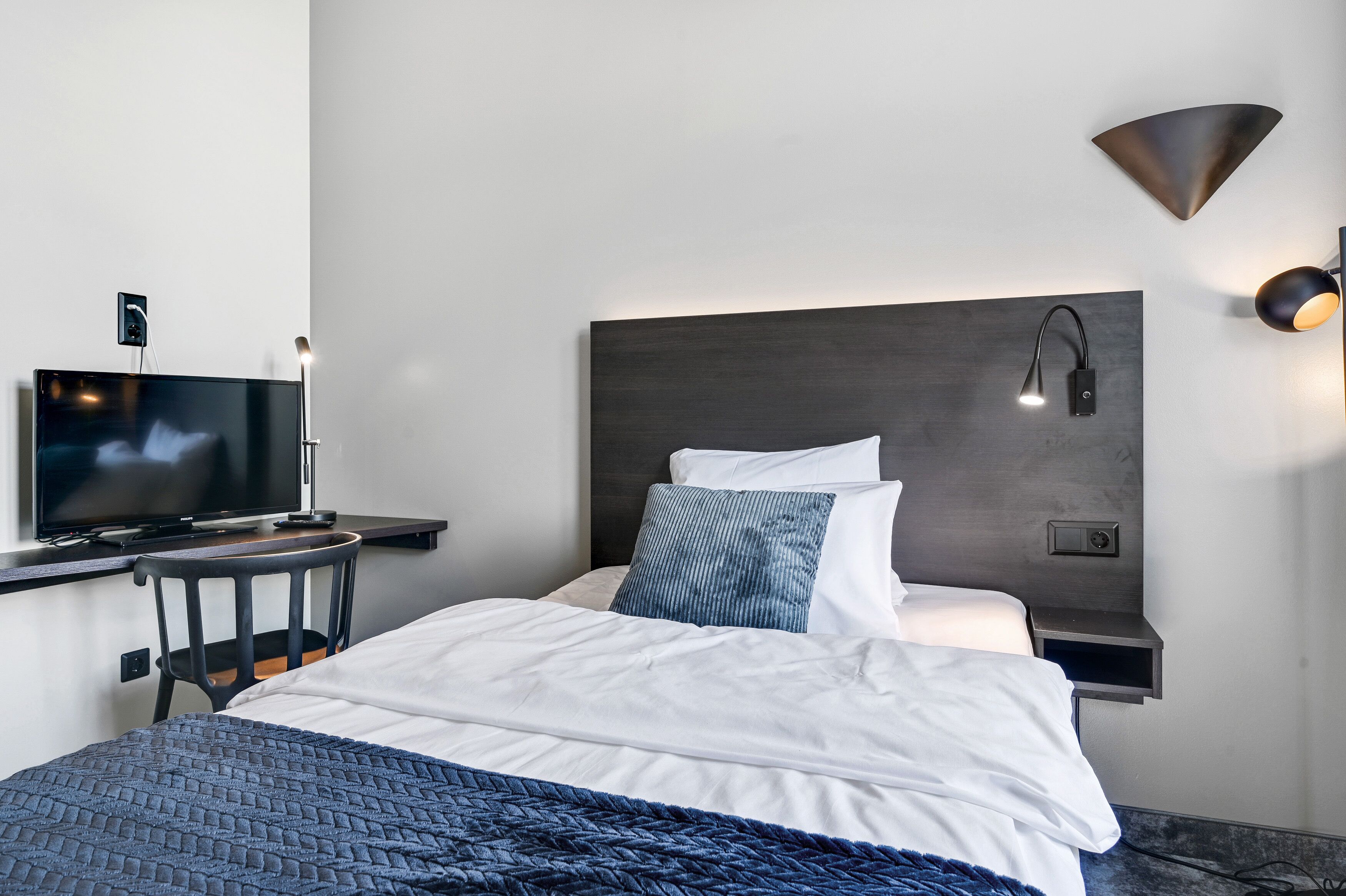 A modern hotel room with a bed, a desk with a television, a wall-mounted lamp, and a power outlet.